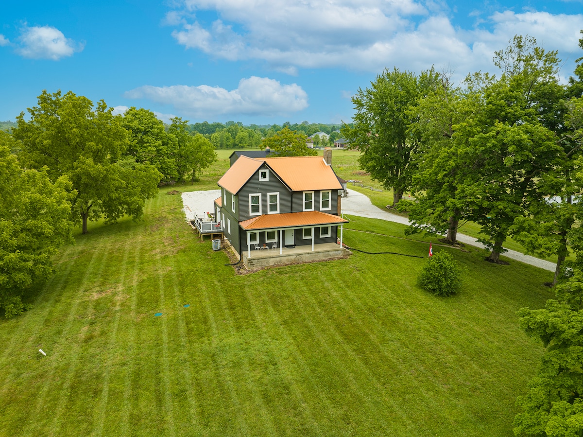 An aerial view of a spacious farmhouse featuring a large green lawn surrounded by mature trees. The structure includes a vibrant orange roof and wide porch. The tranquil rural setting is complemented by the nearby gravel drive and open fields in the background.