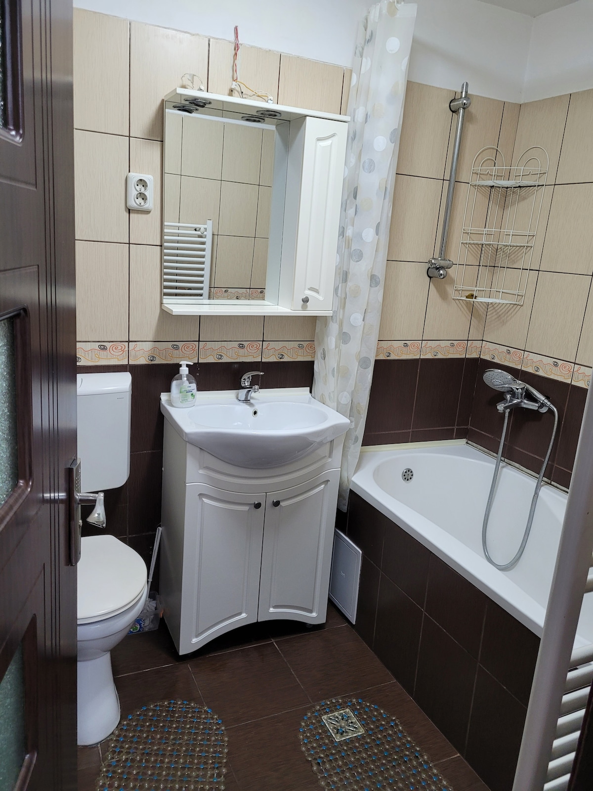 A compact bathroom features a white bathtub with a showerhead, a matching sink with storage beneath, and a stylish mirror above. Neutral-colored tiles create a harmonious appearance, with decorative accents around the space. Soft bath mats are placed near the sink and tub for comfort.