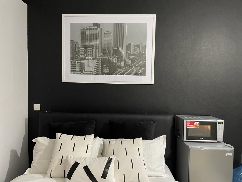 A dark wall serves as the backdrop for a large black-and-white cityscape picture above a dark headboard. The bed is adorned with patterned cushions, while a small microwave is positioned next to it, adding functionality to the sleeping area.
