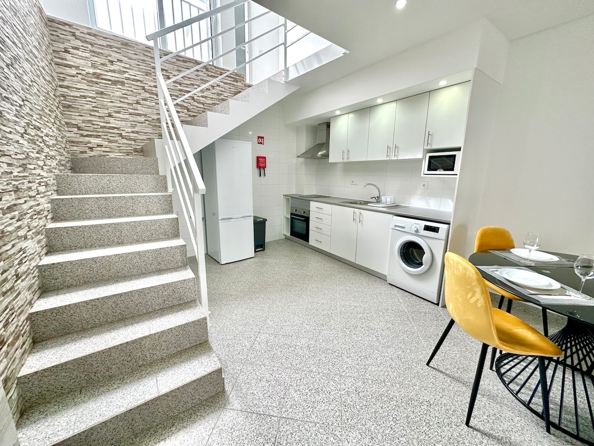 A well-appointed open space suite is presented, featuring a modern kitchen with stainless steel appliances. A dining table with yellow chairs is situated near the kitchen. Stairs lead to an upper level, and natural light illuminates the sleek, neutral-toned decor.