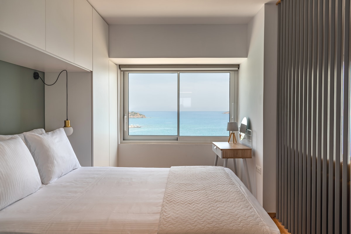 A spacious bedroom is highlighted by a large window offering unobstructed sea views. The bed is neatly made, complemented by a small desk with a lamp nearby. Soft, neutral tones enhance the serene atmosphere of the room, providing a calm retreat.