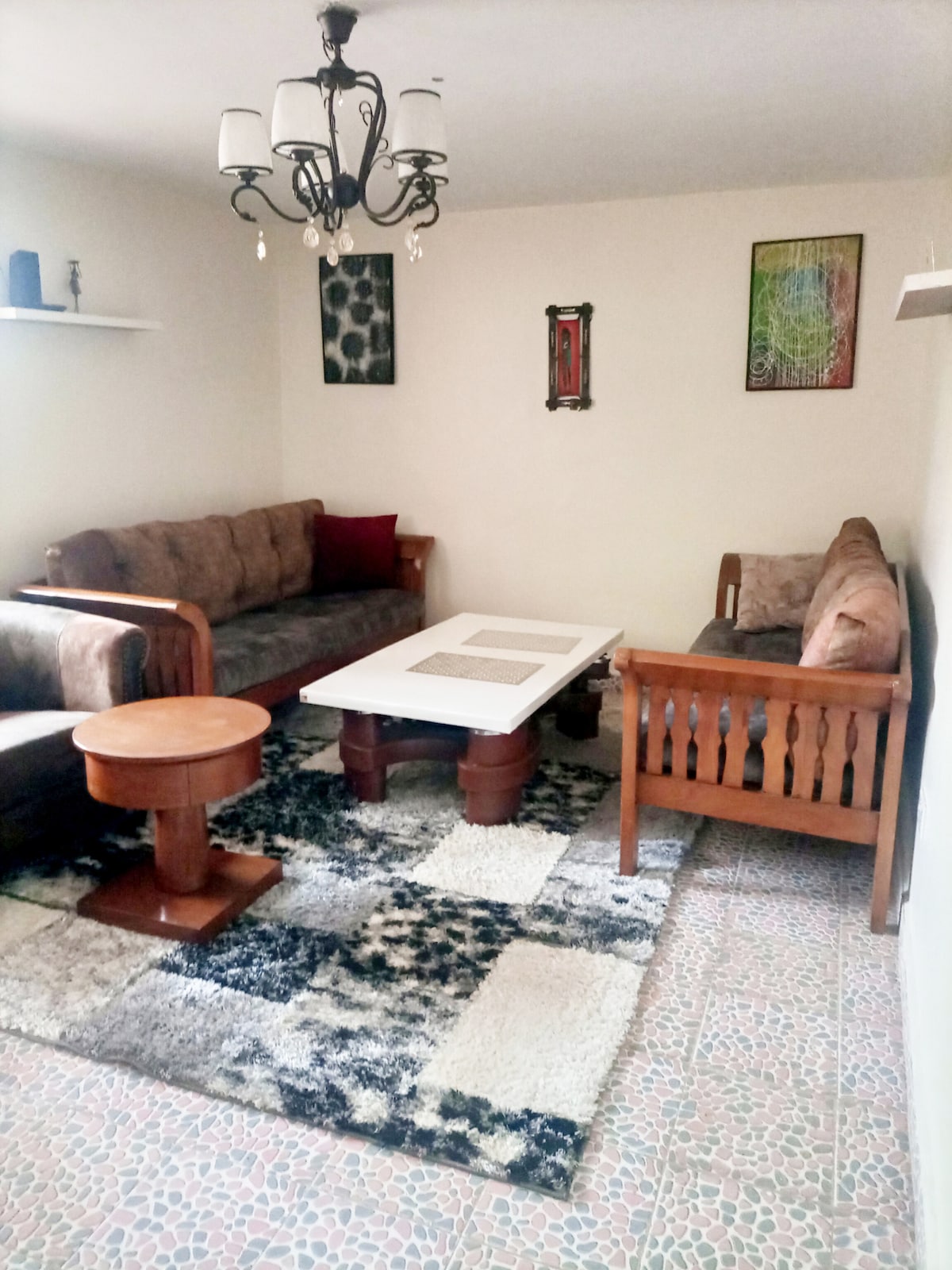 A cozy sitting area is equipped with two cushioned sofas and a wooden bench, surrounded by decorative shelves. A central coffee table rests on a patterned rug, with a chandelier above providing ambient lighting. Brightly colored wall art adds interest to the neutral-toned walls.