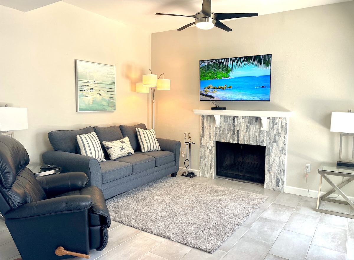 The living room features a gray sofa with decorative pillows, a recliner chair, and a coffee table. A large TV is mounted above a stone fireplace. Soft lighting is provided by floor lamps and ceiling fans, with light gray tiles covering the floor.