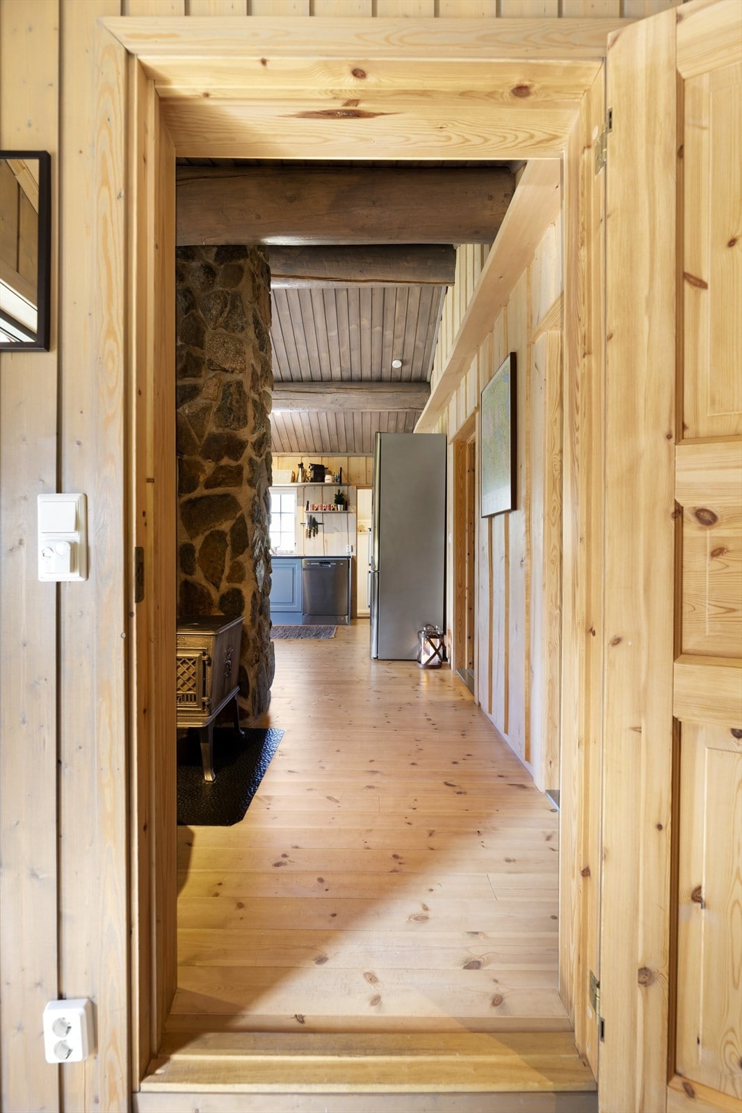 A wooden hallway is framed by light brown paneling, leading to a spacious kitchen area. Features include a stone wall to the left and a glimpse of appliances and cabinetry. Natural light illuminates the wooden floor, enhancing the inviting feel of the cabin.