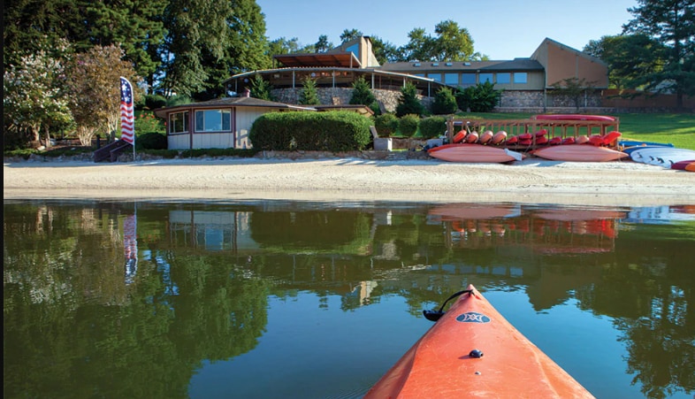 A serene lakeside view is captured, showcasing a sandy beach with calm waters reflecting the surrounding greenery and resort buildings. Several kayaks and paddleboards are lined up along the shoreline, ready for use, while the main building is visible in the background under a clear blue sky.