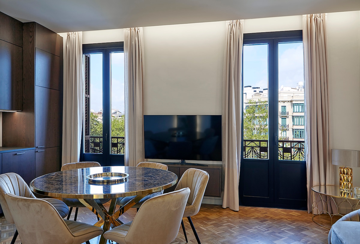 A dining area features a round table with a textured surface, surrounded by upholstered chairs. Large windows allow natural light to enter, revealing a view of the surrounding buildings. A flat-screen television is mounted on the wall, contributing to the modern decor.