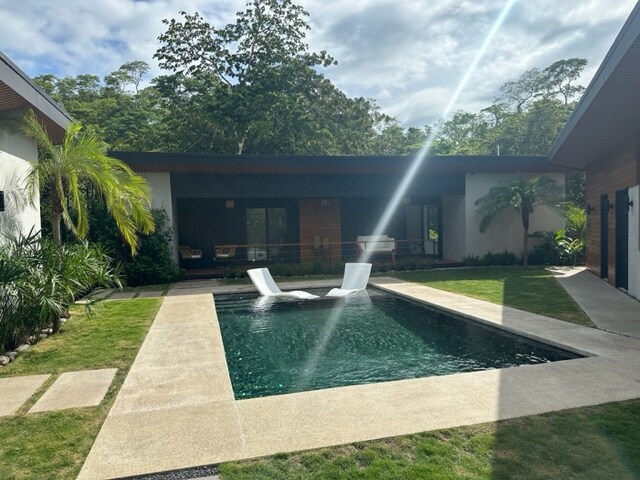 A modern outdoor area features a refreshing pool with sleek lounge chairs positioned on one side. Lush greenery surrounds the space, with well-maintained grass and tropical plants providing a serene backdrop. Large glass doors are visible, connecting the area to the interior.