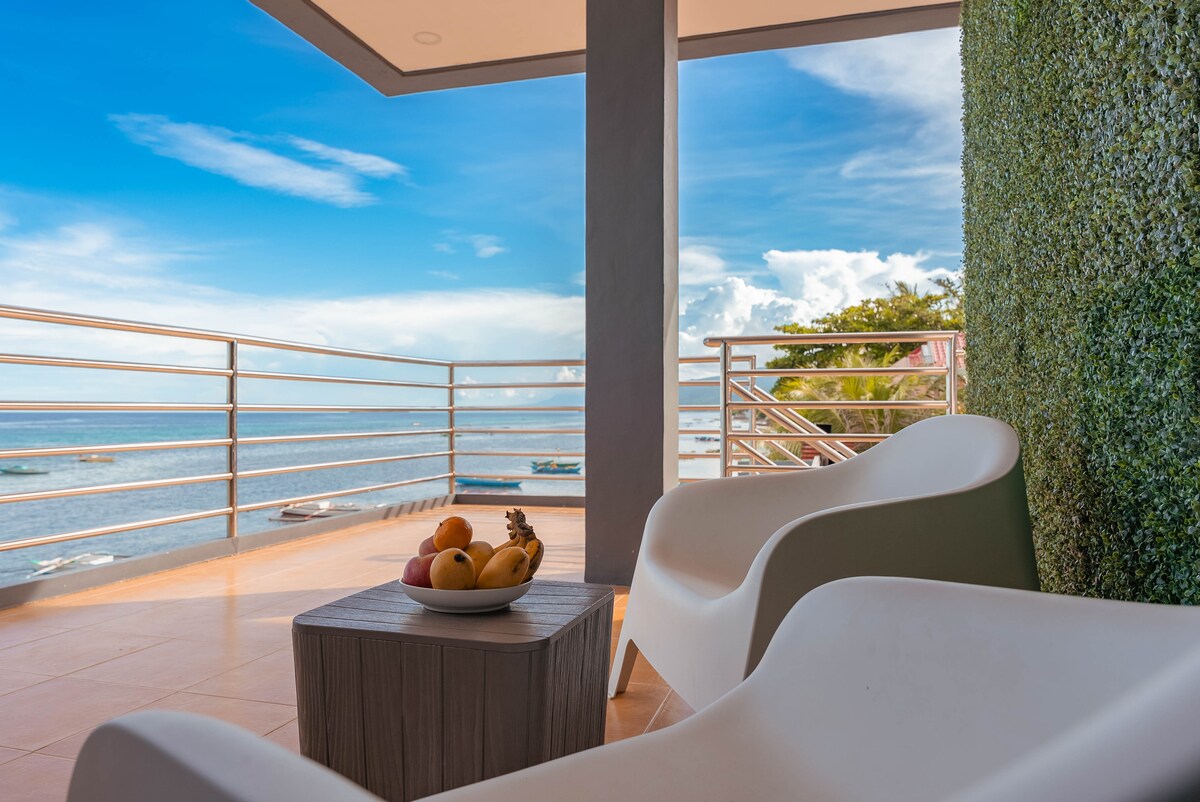 A comfortable seating area features sleek white chairs arranged with a small wooden table adorned with a bowl of assorted fruits. The scene includes a partial view of the sea and clear sky, framed by a railing along the balcony.
