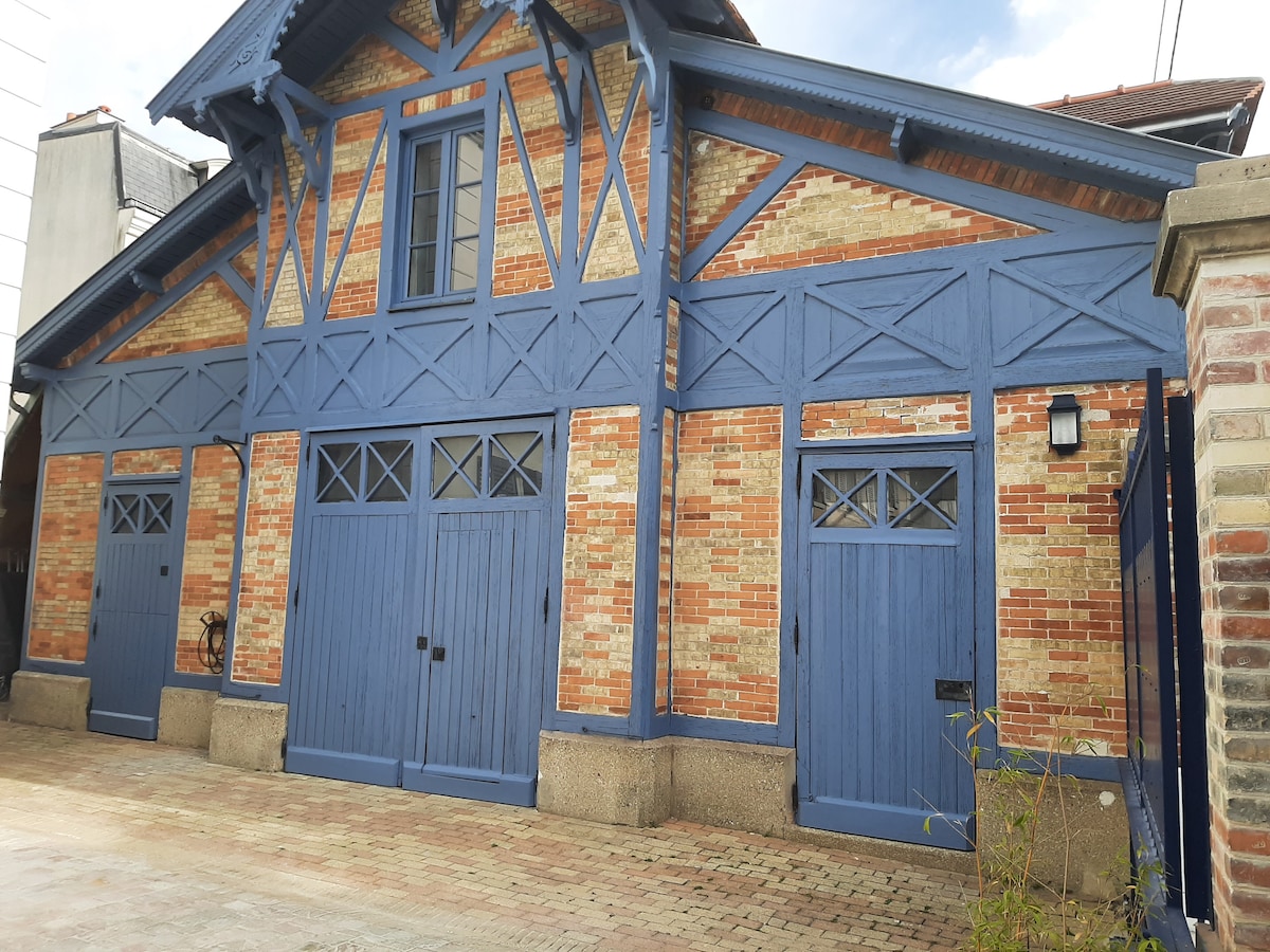 The exterior of a charming renovated stable is presented, featuring distinctive blue wooden details and traditional brickwork. The façade includes several doors with decorative grilles and geometric window patterns, surrounded by a paved courtyard that leads to a gated entrance.