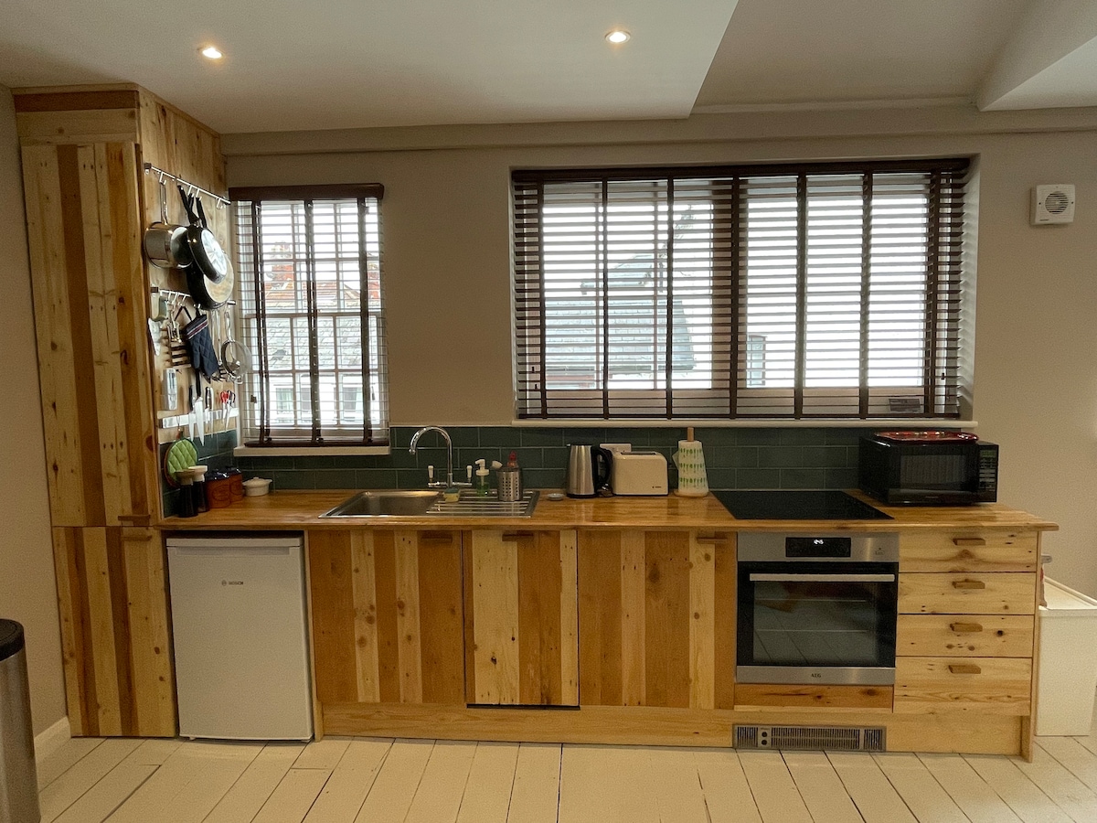 The kitchen area features wooden cabinetry with a natural finish and modern appliances, including a refrigerator, microwave, and oven. Ample natural light enters through multiple windows with wooden blinds, highlighting the simple yet functional design of the space.