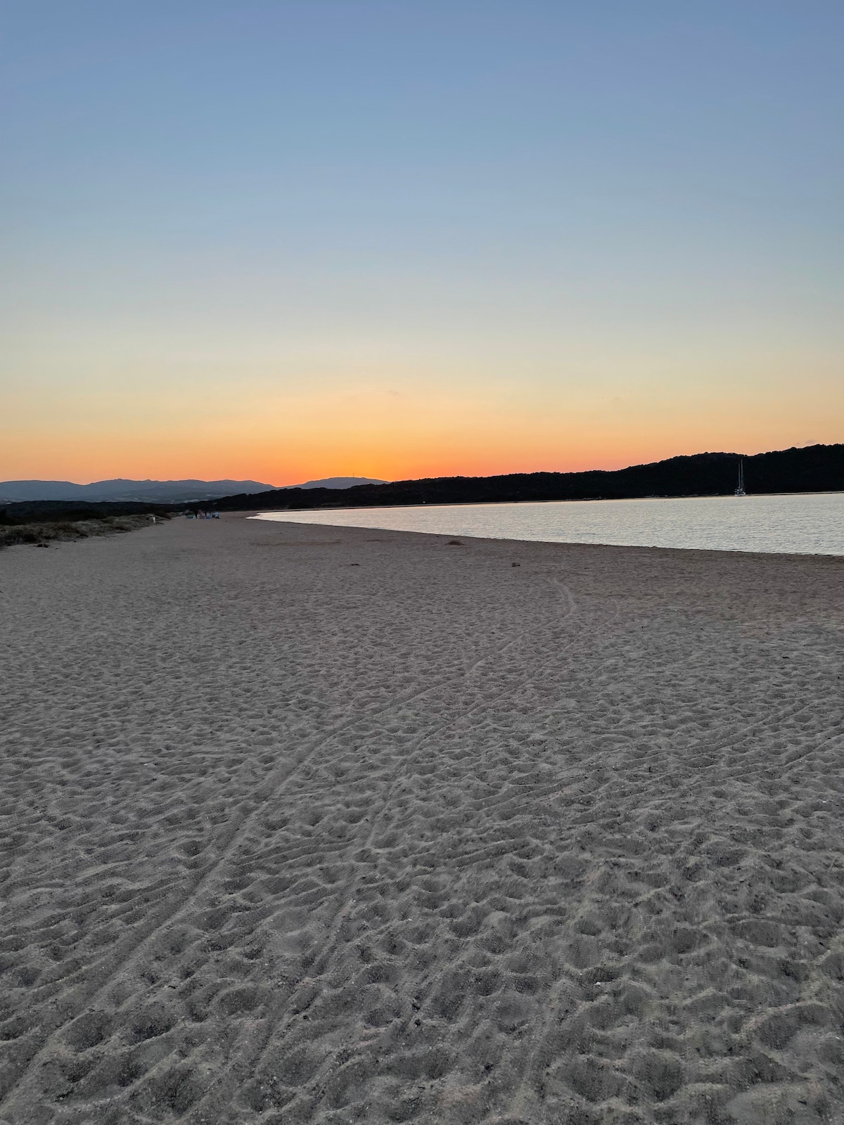 A serene beachscape is depicted at sunset, showcasing soft sandy shores that stretch into the distance. The sky transitions from warm hues of orange to cooler shades, while gentle waves lap at the edge of the water, reflecting the colors of the sunset.