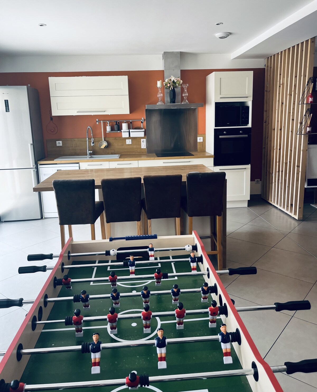 A foosball table occupies the foreground, featuring brightly colored player figures. In the background, a modern kitchen showcases sleek cabinetry, a stainless steel refrigerator, and a four-seat bar area. Natural light illuminates the space, enhancing the warm hues of the walls.