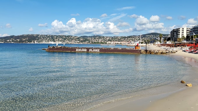 A serene beach scene is depicted, featuring clear turquoise waters that gently lap against the sandy shore. In the distance, a promenade is visible with various buildings, while a breakwater extends into the water, marked with signage. Fluffy clouds float above, adding to the tranquil atmosphere.