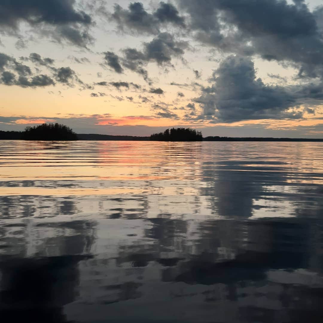 A serene lake scene is captured at sunset, featuring gentle ripples reflecting hues of orange, pink, and grey in the sky. Small islands are visible in the distance, contributing to the tranquil ambiance of the water's surface.