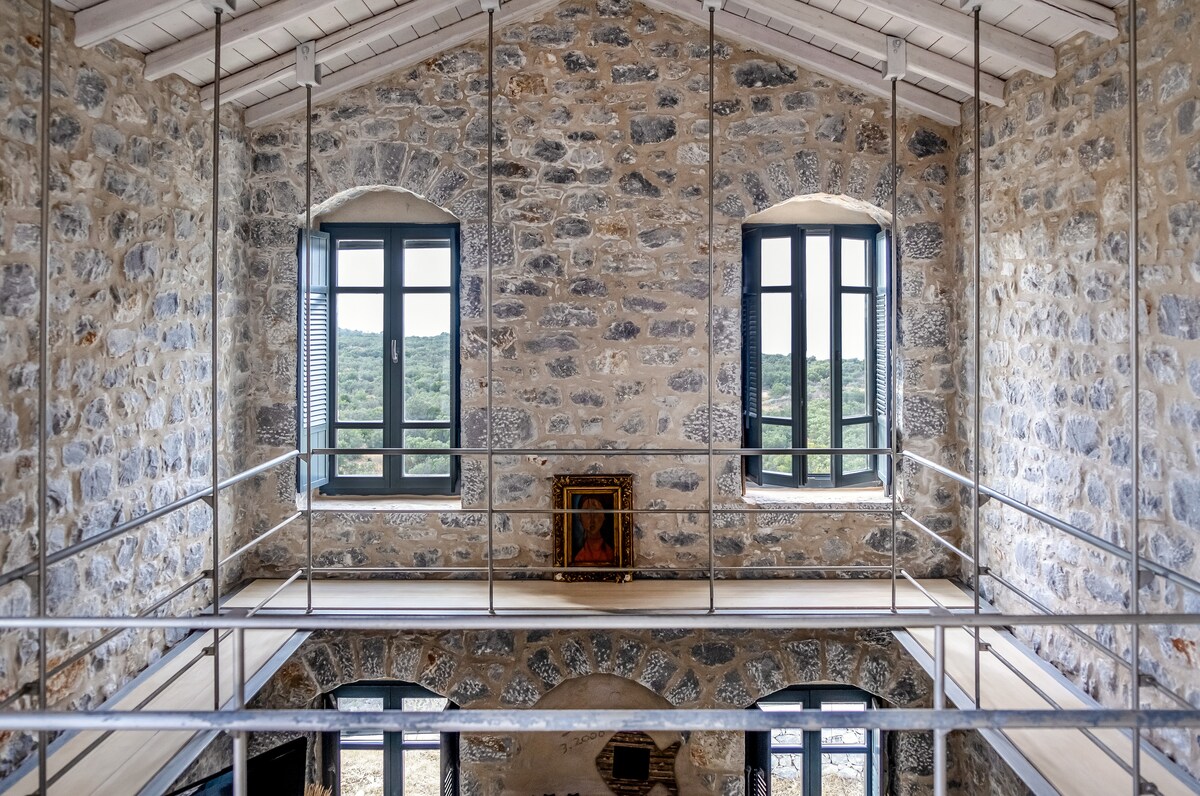 The interior features exposed stone walls, showcasing the home's historical character. Large windows on either side allow natural light to fill the space, while a railing outlines a second-floor balcony that overlooks the scenery, adding an airy and open feel.
