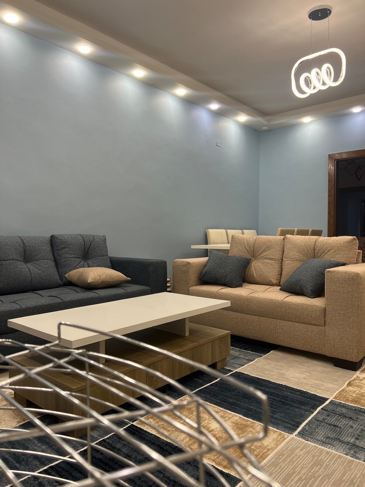 A cozy living area features two seating arrangements: a dark grey couch and a light beige sofa with accented cushions. A central coffee table stands between them, surrounded by a patterned area rug. Soft overhead lighting creates a warm ambiance throughout the space.