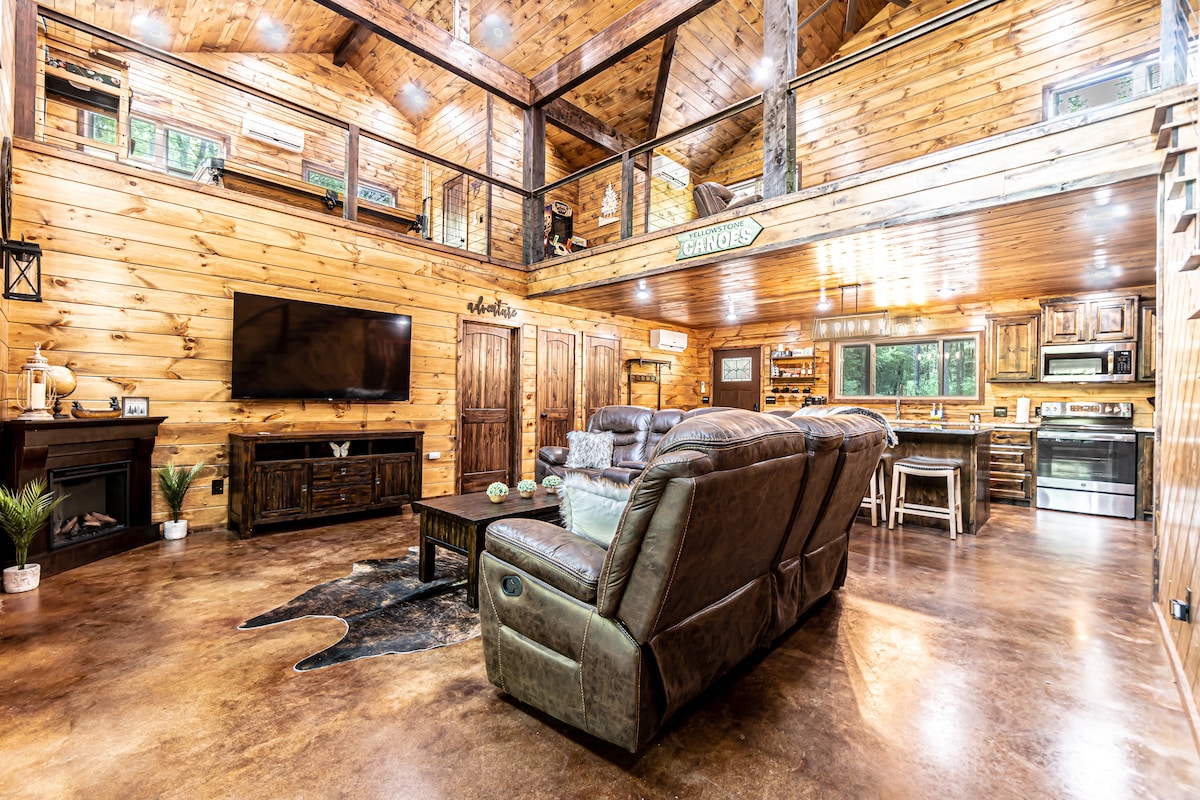 A spacious living area features wooden walls and ceiling beams, emphasizing a cozy cabin ambiance. Comfortable seating is arranged around a coffee table. A large flat-screen TV is mounted on the wall, and a fully equipped kitchen is visible in the background.