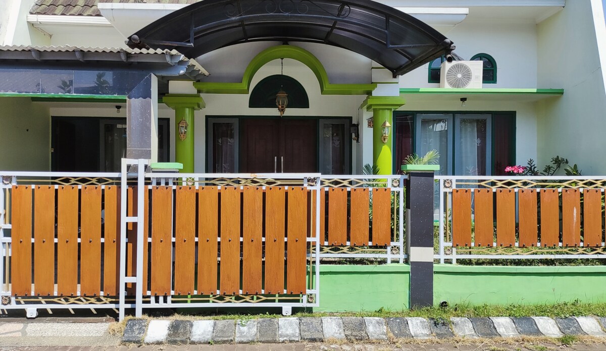 The exterior of the house features a welcoming entrance with a curved roof overhang. A decorative wooden gate, painted in earthy tones, complements the vibrant green accents of the facade. Lush plants are visible in the garden area, enhancing the inviting atmosphere.