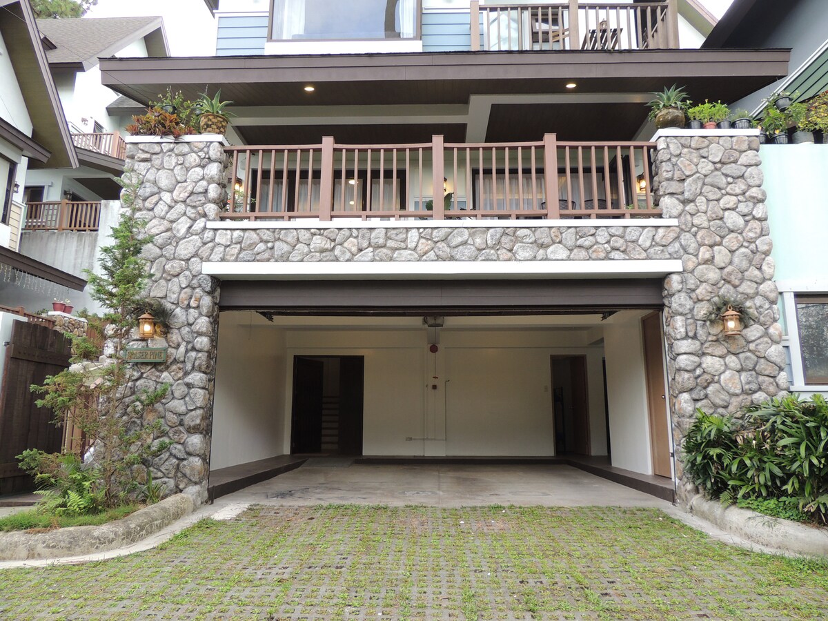 The exterior view showcases a stone-clad garage area designed to accommodate two cars. A spacious balcony is positioned above, framed by wooden railings. Lush greenery is displayed along the sides, contributing to the welcoming appearance of the villa.