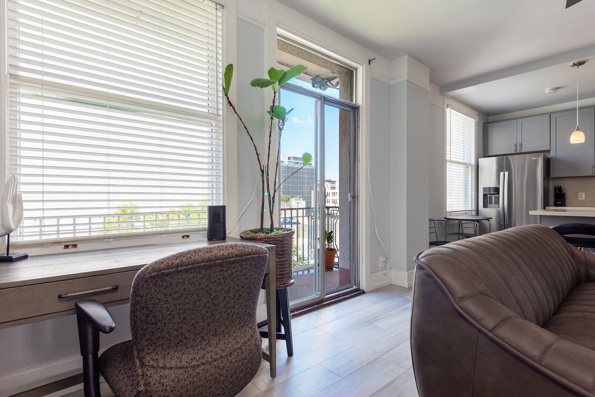 A well-lit living area features a modern seating arrangement and a workspace with a comfortable chair. Large windows with blinds allow natural light, and a potted plant adds a touch of greenery. A sliding glass door leads to a small balcony, enhancing the indoor-outdoor connection.