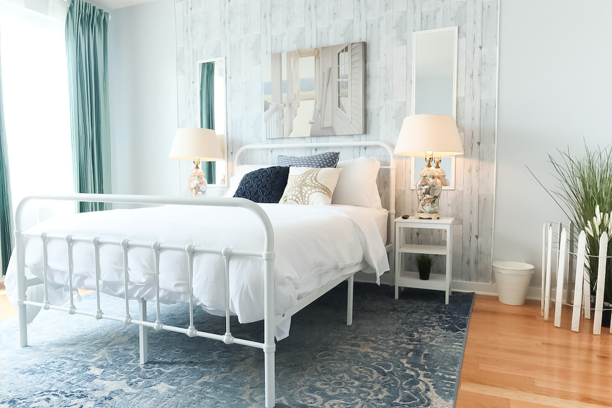 A bright bedroom showcases a white metal bed frame with a spacious bed covered in white linens. Two bedside lamps illuminate the room, complemented by mirrored accents. A blue area rug adds warmth, while large windows allow natural light to fill the space, framed by lush green curtains.