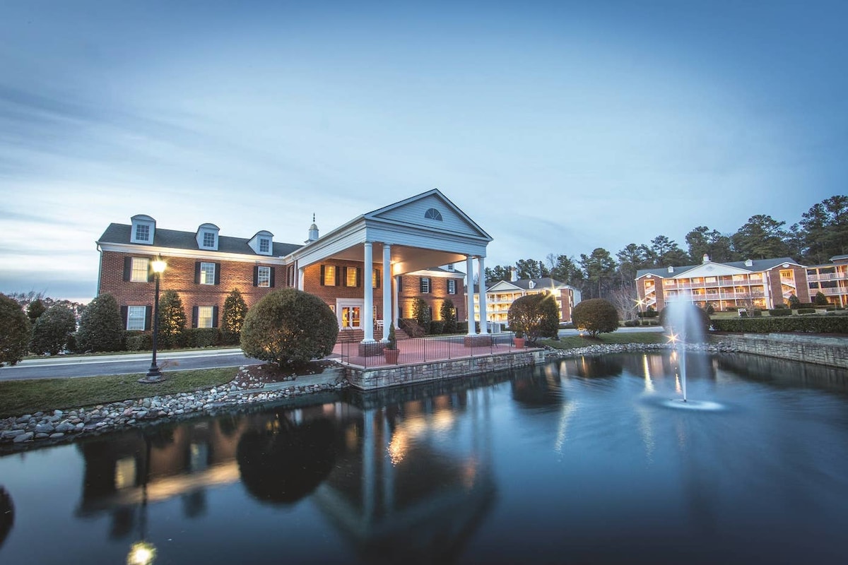 The resort's exterior features a colonial-style building with a prominent portico and well-maintained landscaping. A serene pond reflects the architecture, with a fountain gently flowing, enhancing the peaceful ambiance of the surroundings.