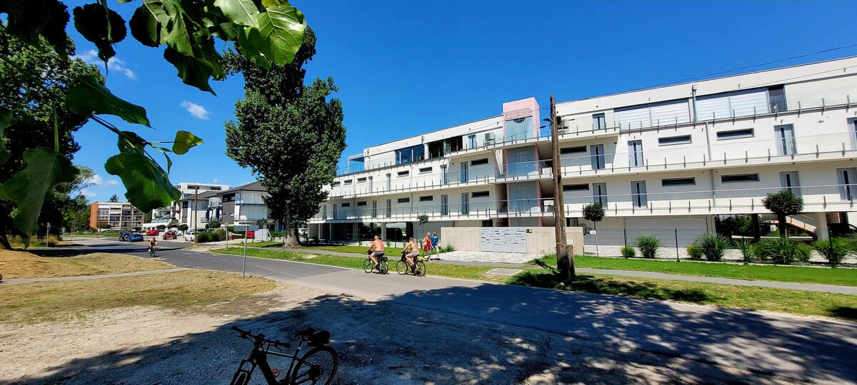 A modern apartment building is visible across a landscaped area, with pathways leading towards it. Some cyclists are seen riding along the path, while greenery and trees provide shade in the foreground. The sky is bright and clear, contributing to an open atmosphere.