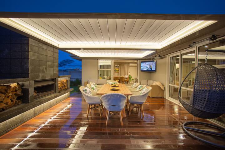Elegant outdoor dining area with a wooden table, cushioned chairs, a hanging chair, and a large fireplace under a lit pergola