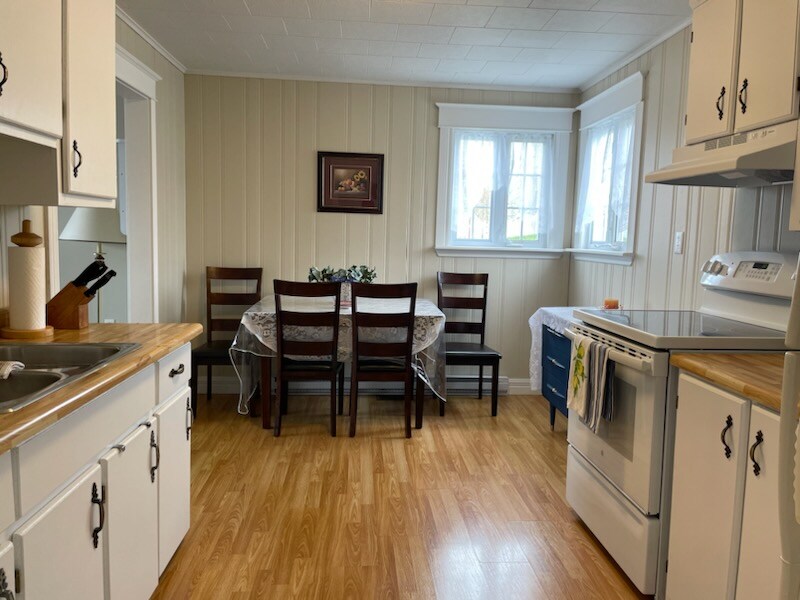 A bright kitchen space is visible with white cabinetry and wooden countertops. A round table is surrounded by six dark wooden chairs, positioned near two windows that allow natural light to fill the room. A stove and sink are also present in the area.