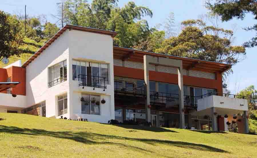 The exterior of the house is displayed, featuring two stories with large glass windows and balconies. A gentle slope of green grass leads to the building, while trees are visible in the background, enhancing the natural setting.