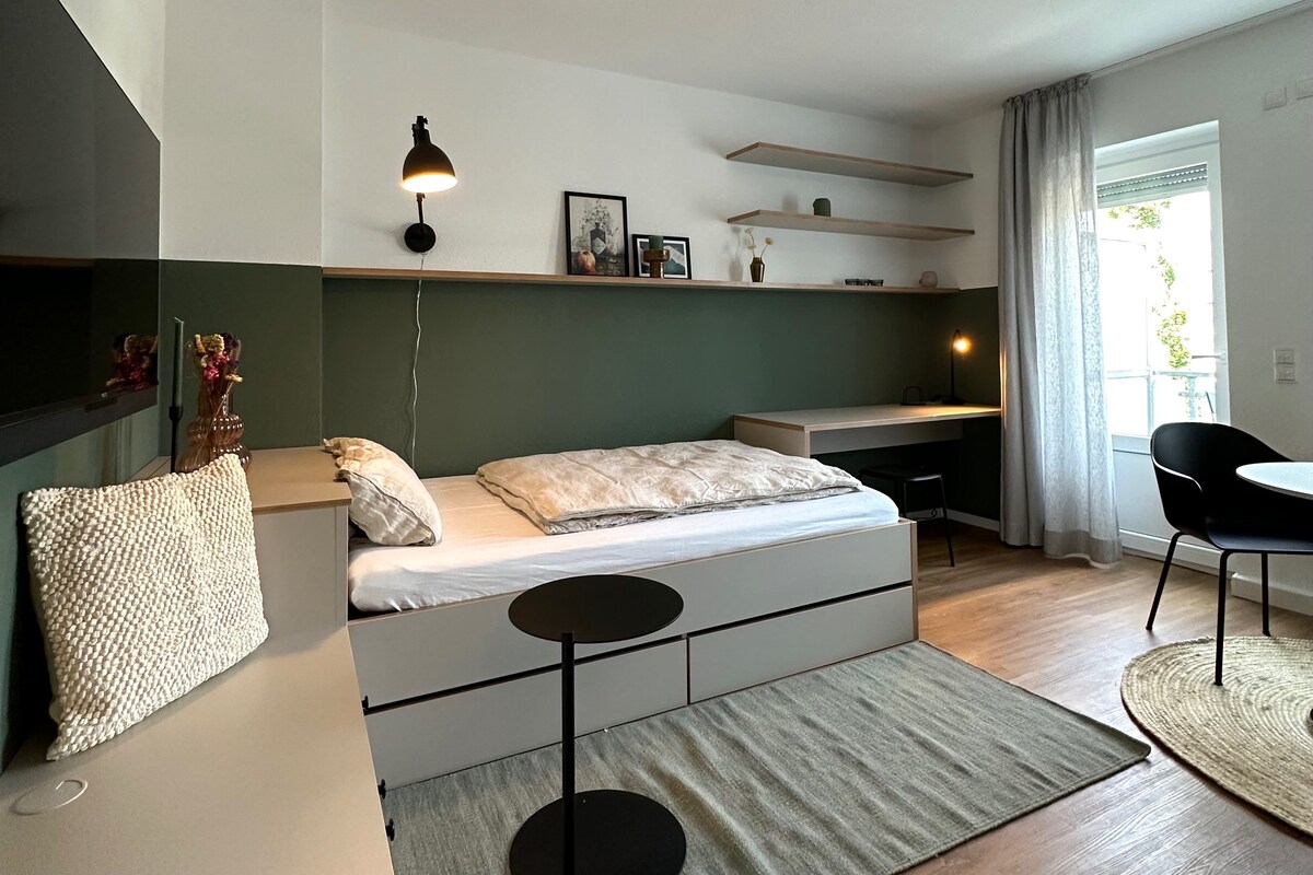 A spacious studio apartment features a comfortable bed with a light-colored comforter, complemented by a neutral rug. Shelves are mounted on the deep green wall, and a small black table sits beside the bed. A desk and chair provide a dedicated work area, with natural light entering from a large window.