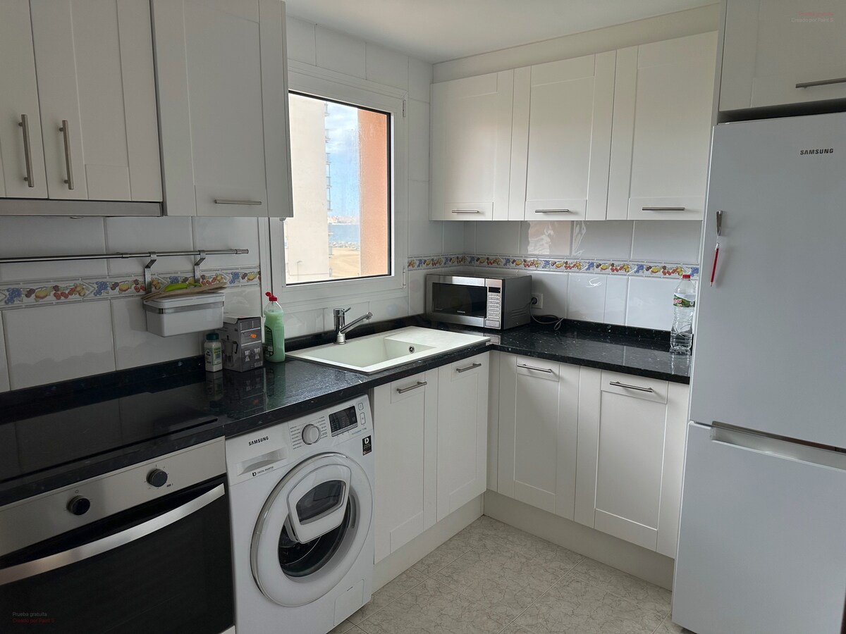 The kitchen features sleek white cabinetry and dark countertops. An oven, washing machine, microwave, and refrigerator are present, providing essential appliances. A window allows natural light to enter, enhancing the functional space.