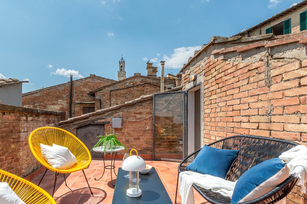 A charming rooftop terrace featuring woven seating arrangements, highlighted by a coffee table and a decorative lamp. Sunlight fills the space, illuminating the brick walls and rooftops of Siena, with a distant view of the tower visible against a clear blue sky.