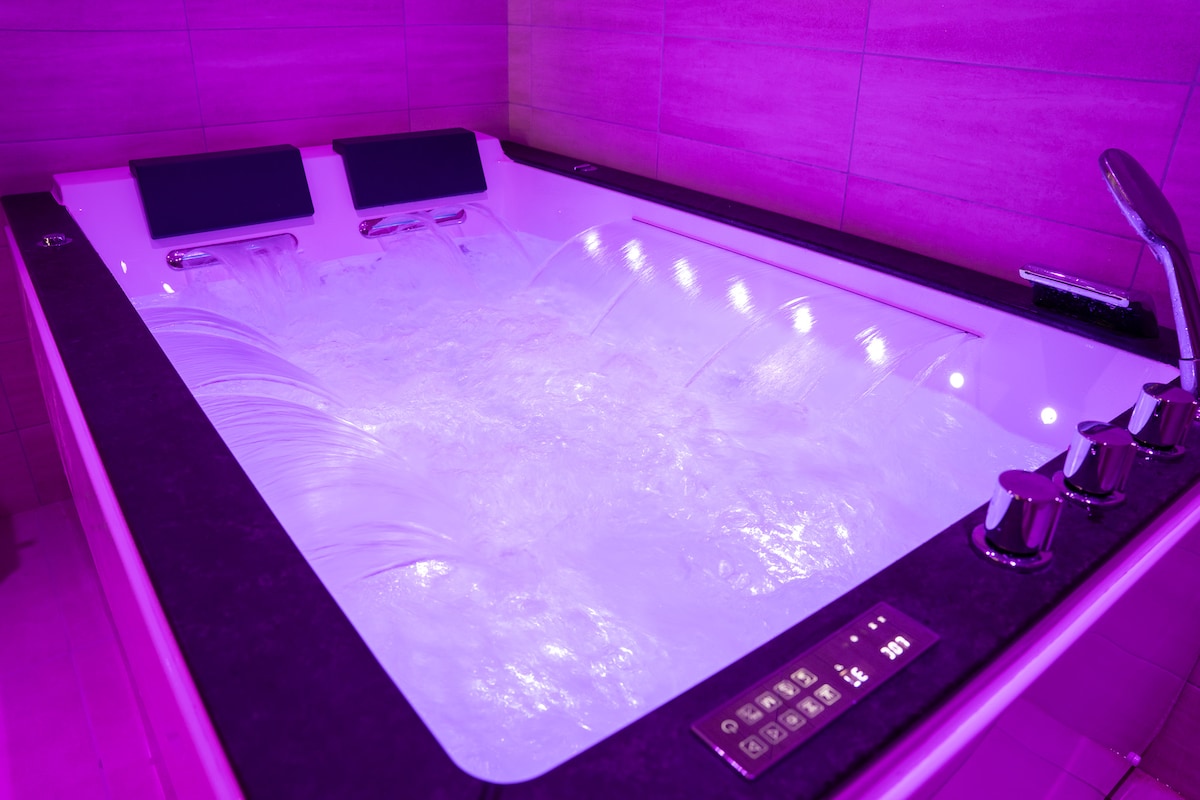 A spacious jacuzzi is featured, illuminated by soft LED lights in a soothing purple hue. The tub includes ergonomic seating for two and controls for various water features, all set against a backdrop of elegant, textured tiles.