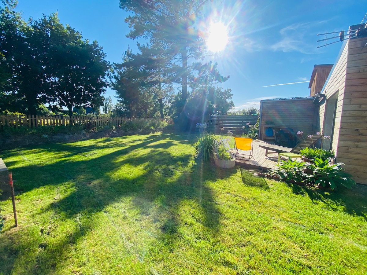 A spacious garden area is illuminated by bright sunlight, featuring well-maintained grass and diverse greenery. A cozy seating area with a vibrant chair and wooden deck invites relaxation. Trees create gentle shadows across the lawn, enhancing the peaceful outdoor setting.