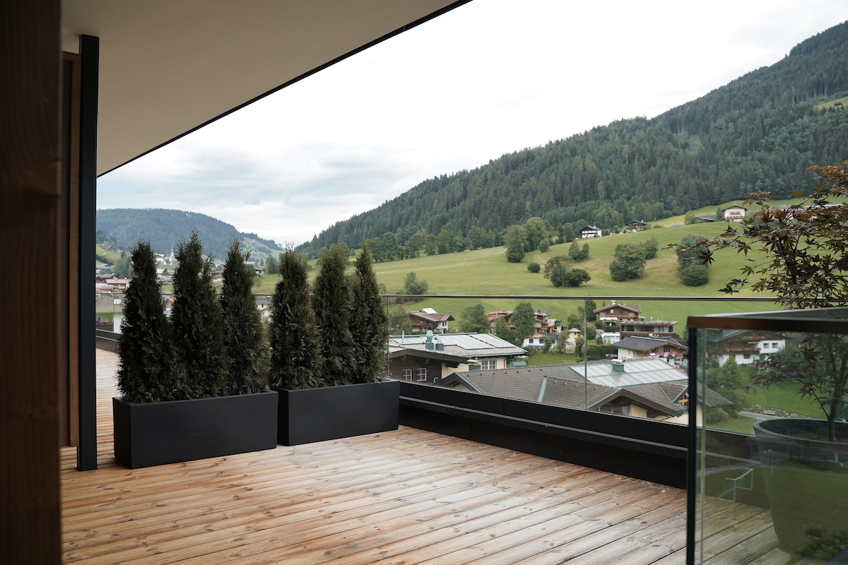 A spacious terrace features wooden flooring and potted evergreen plants, offering a view of the surrounding hills and greenery. The landscape includes scattered residential buildings, with a backdrop of lush mountains under a cloudy sky.