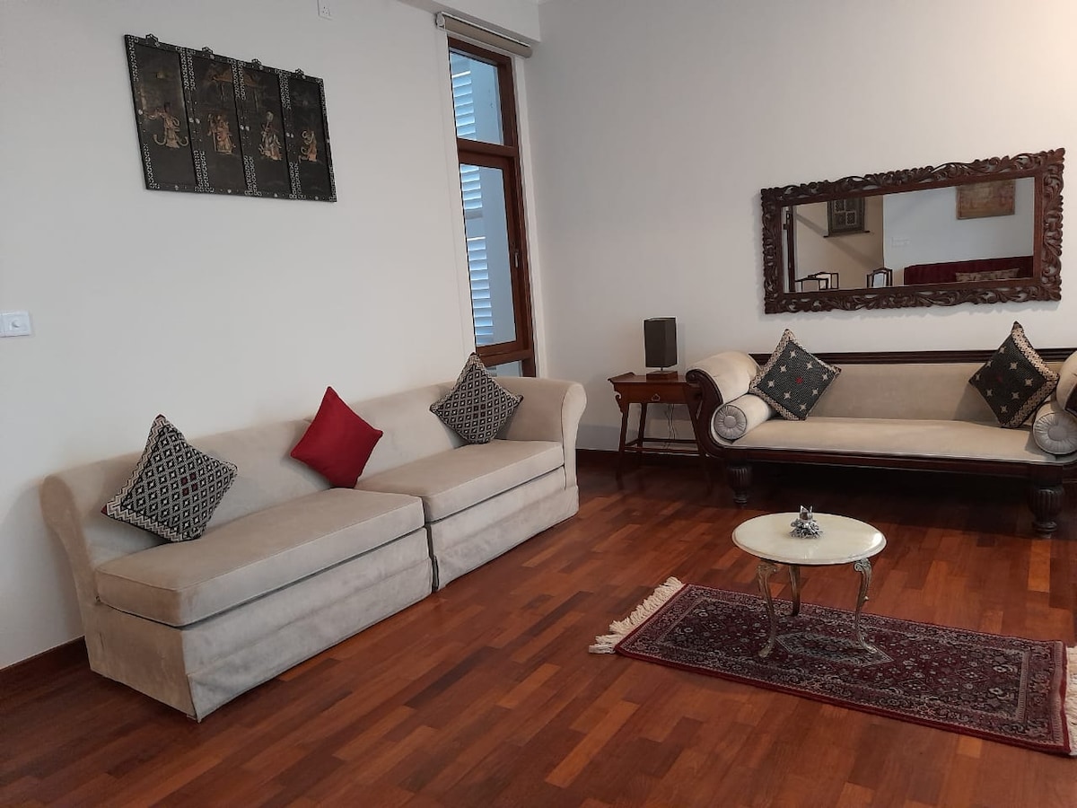 A spacious living area features two elegant sofas adorned with decorative cushions. A small round table sits on a traditional rug, complementing the wooden flooring. A large mirror and wall art add depth to the room, while natural light filters in through the windows.