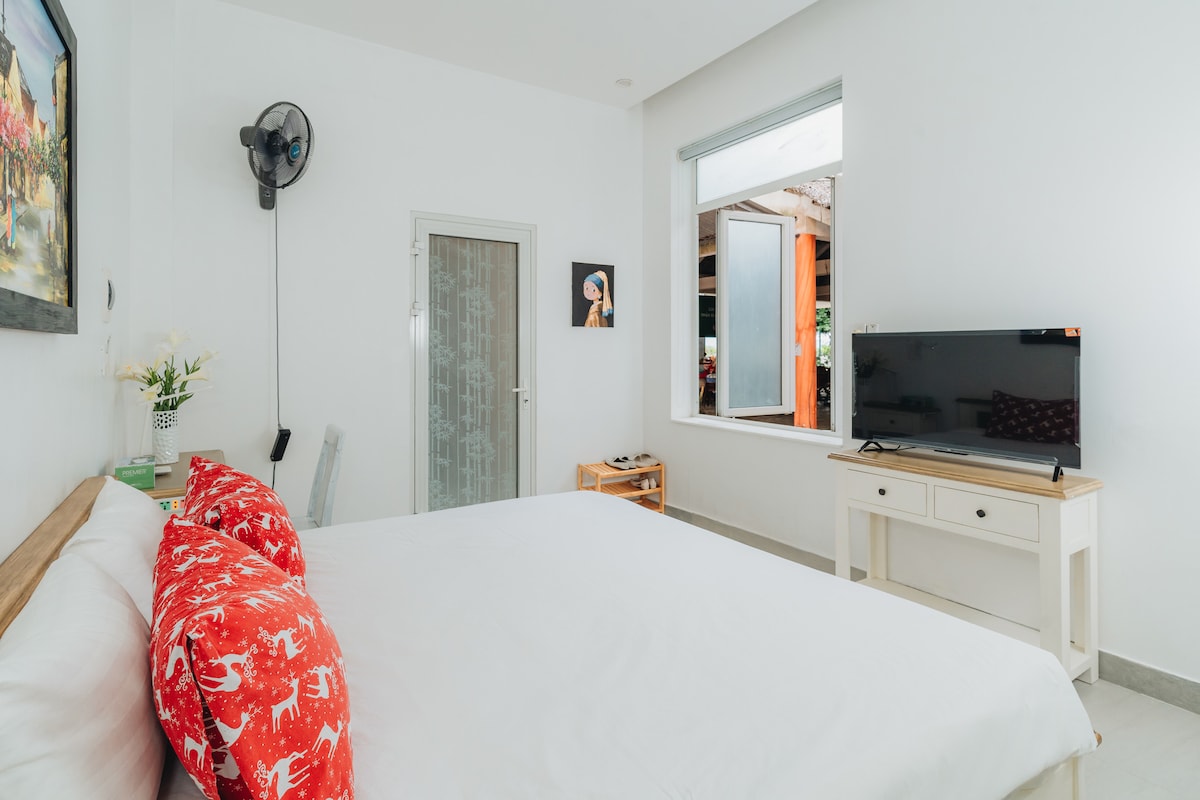 A spacious sleeping area features a comfortable bed with a crisp white duvet and vibrant red accent pillows. A flat-screen television is positioned on a simple white console. Natural light enters through a large window, illuminating the neutral-colored walls.
