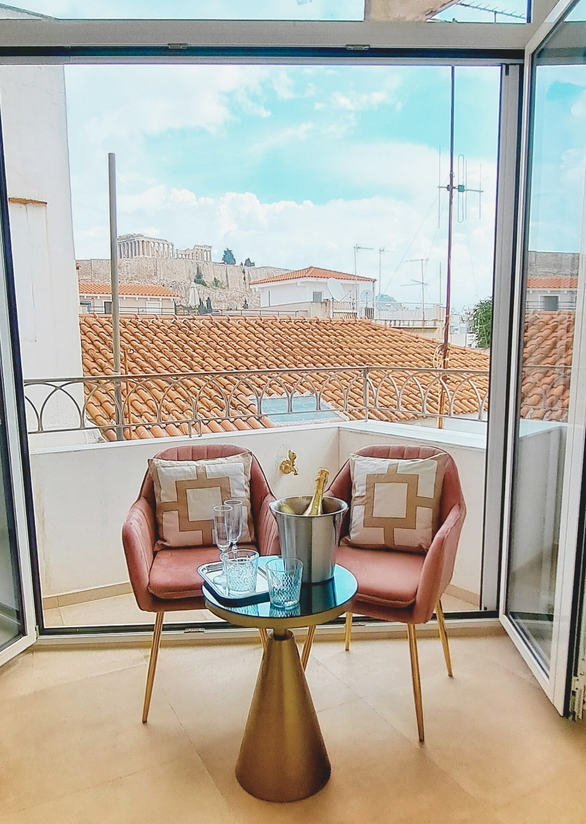 A bright balcony features two pink chairs and a small round table adorned with a silver bucket and glassware. A view of the Acropolis can be seen in the distance, with terracotta rooftops framing the scene beyond the glass doors.