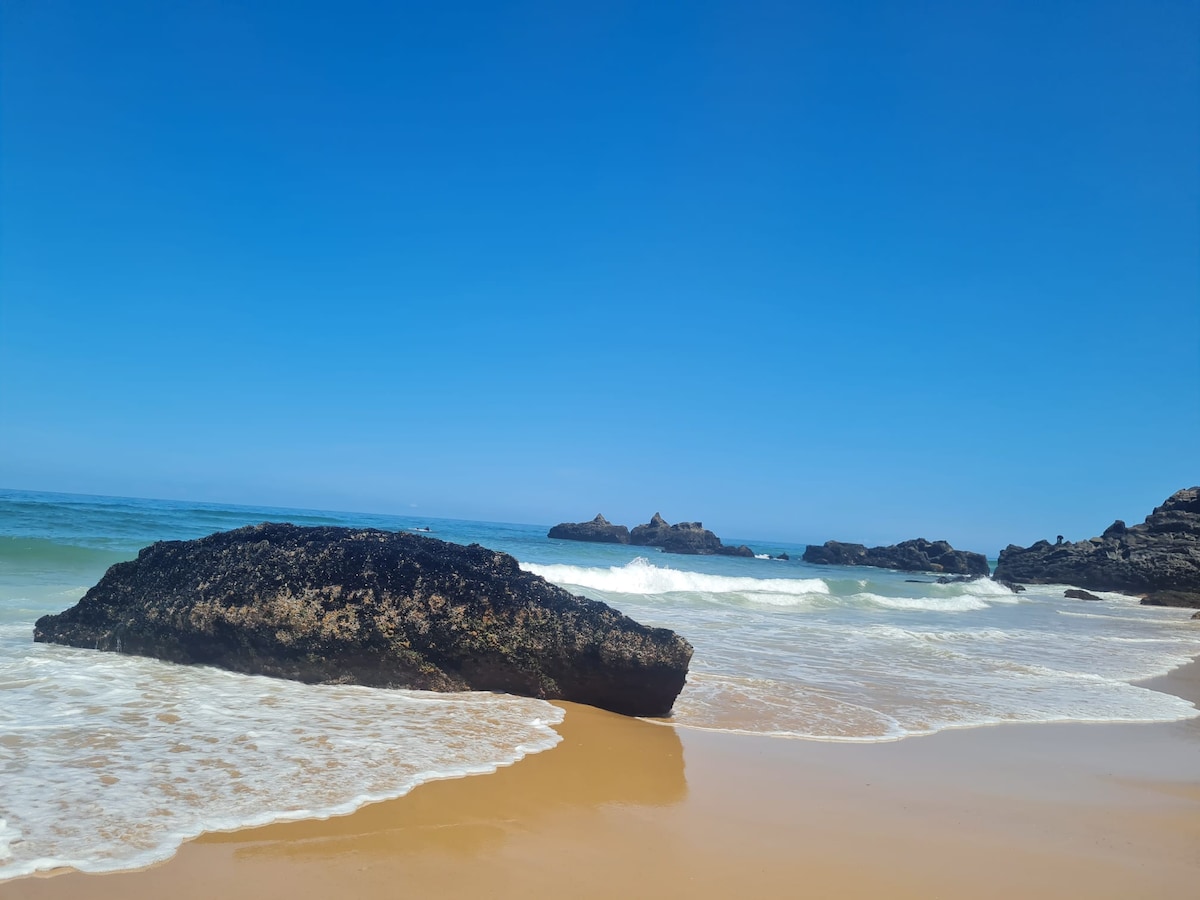A serene beach scene is depicted, with gentle waves lapping at the shoreline. Large, textured rocks are positioned on the sand, contrasting with the clear blue sky and ocean. The horizon is unobstructed, creating a tranquil atmosphere.