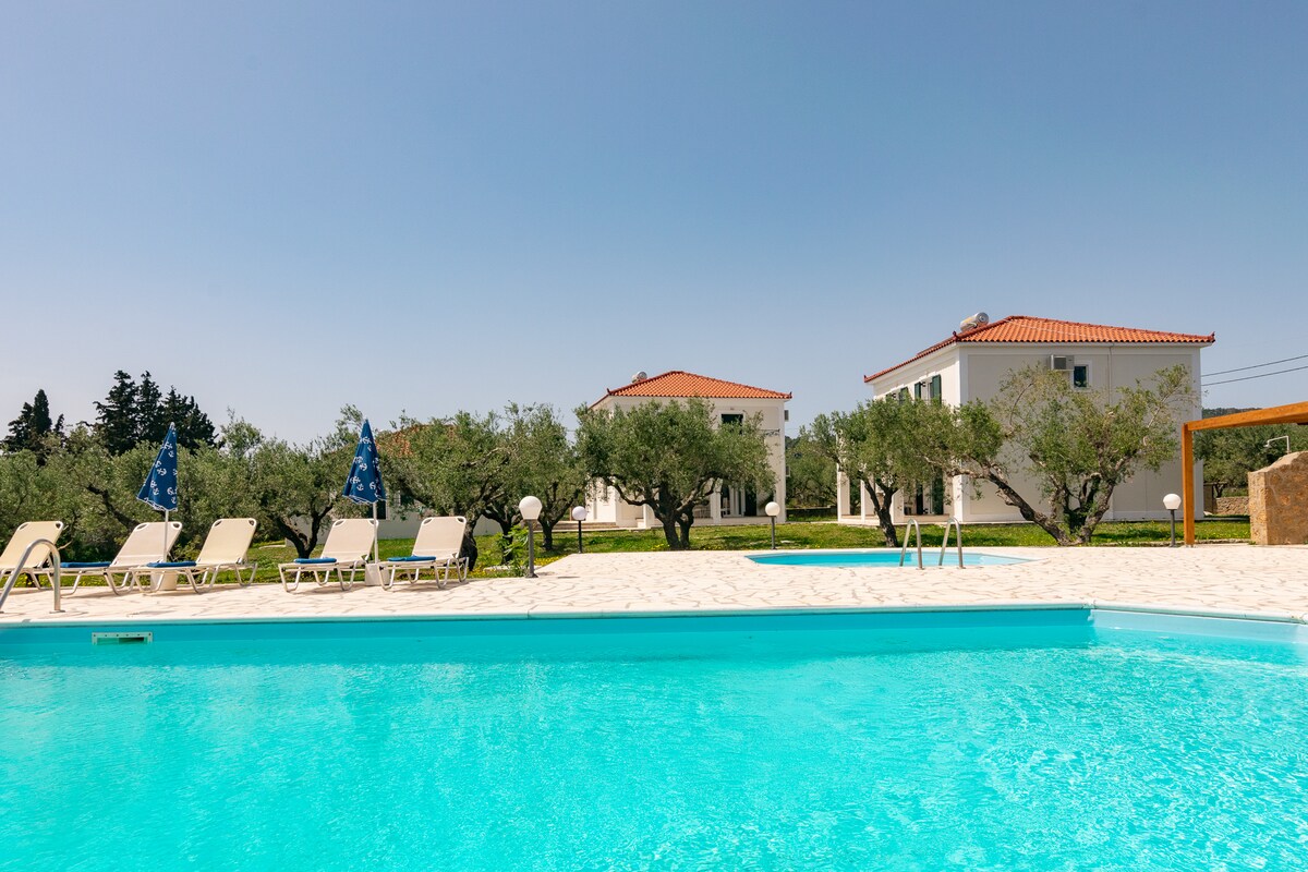 A clear blue swimming pool reflects the sunlight, surrounded by olive trees and sun loungers. Two white villas are positioned in the background, emphasizing the serene landscape. The pool area is paved and features a gentle gradient leading to the water.