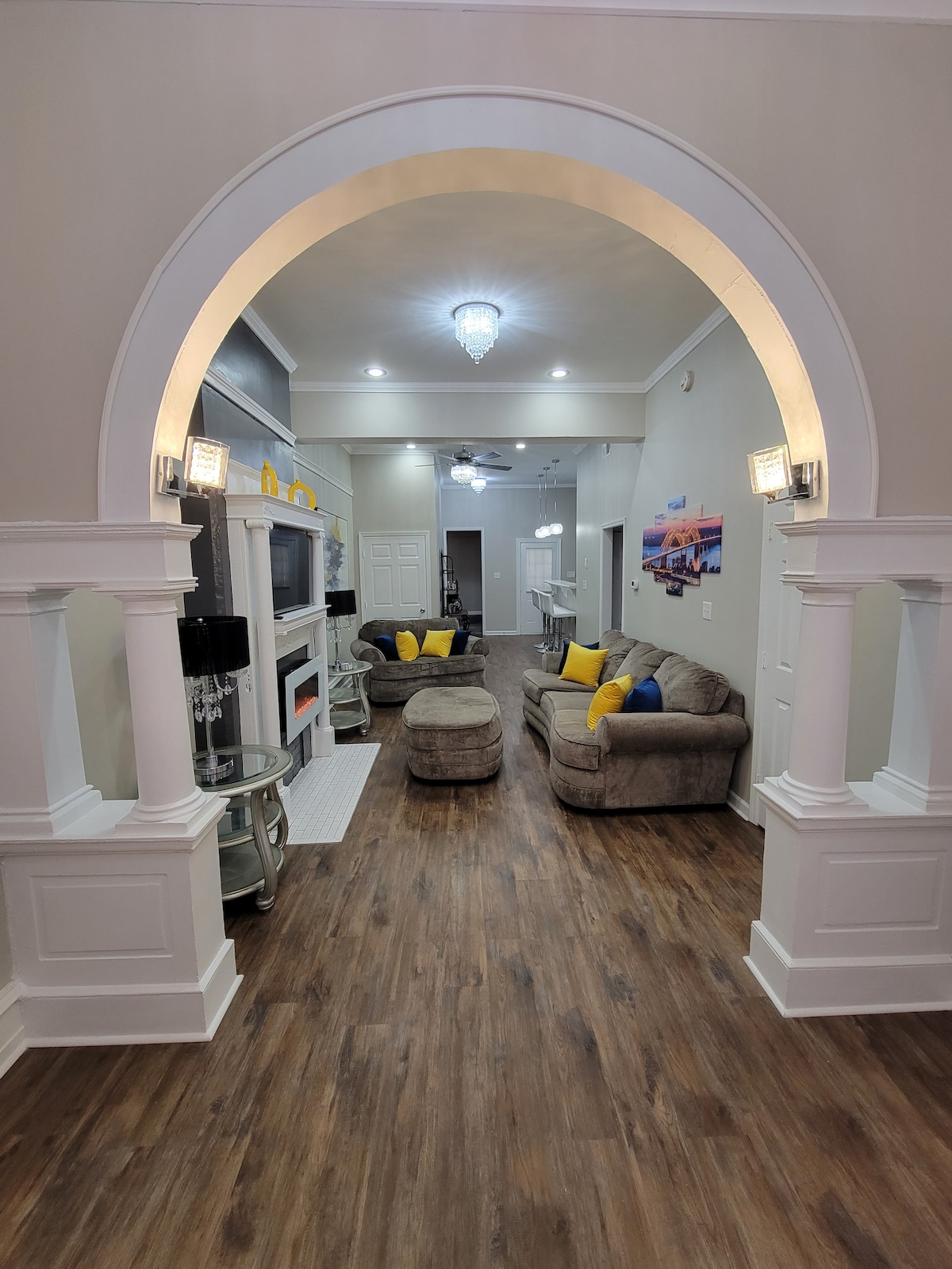 An entryway framed by an elegant arch leads into a spacious living room. Two comfortable sofas adorned with yellow cushions are arranged around a central space. Soft overhead lighting illuminates the wooden floors, enhancing the inviting ambiance of the area.