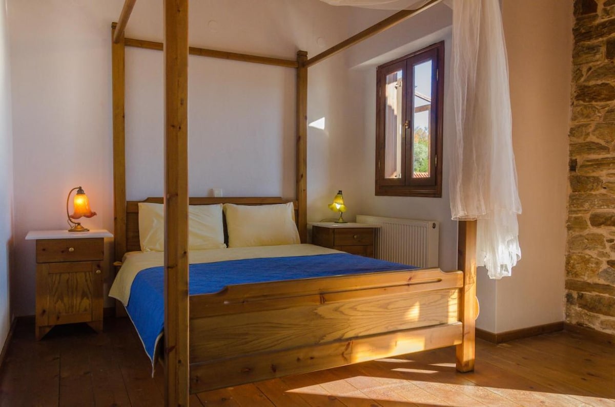 A wooden bed frame is featured, complemented by soft, neutral bedding. Two bedside tables with lamps provide gentle illumination. A window allows natural light to enter, highlighting the stone accent wall and hardwood flooring in the spacious room.