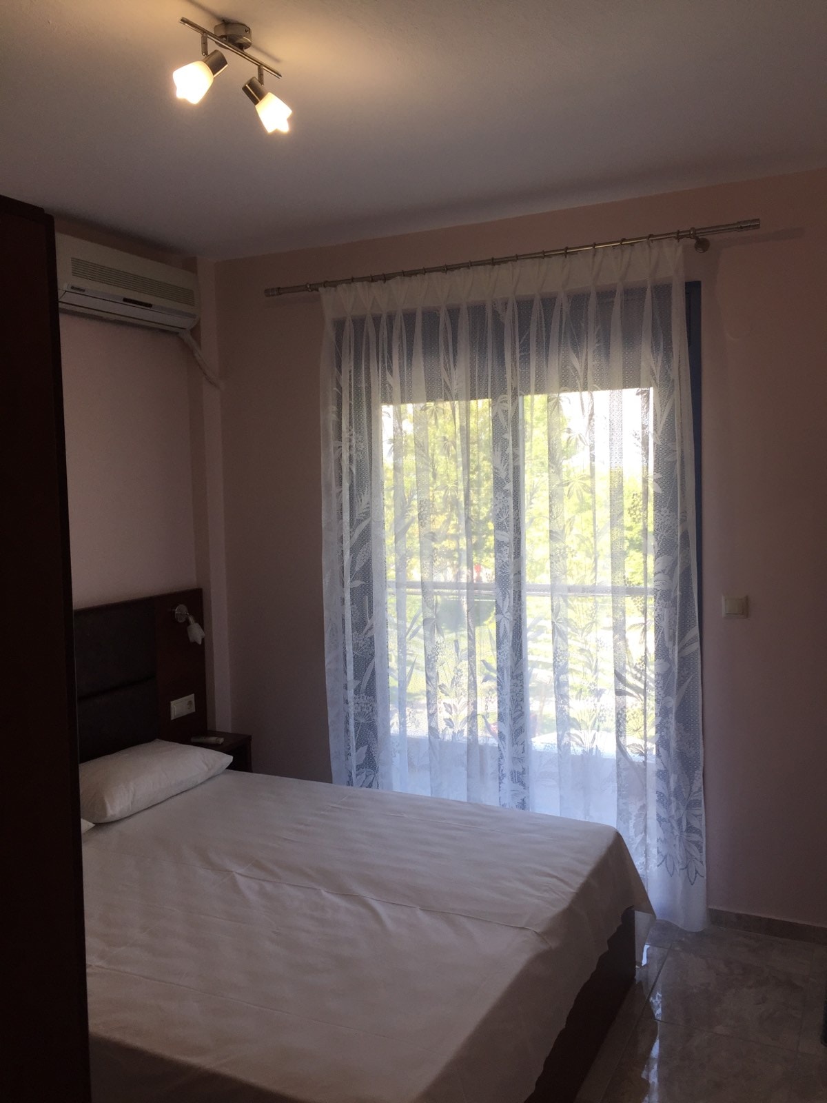 A bright sleeping area features a spacious bed against a soft pink wall. Natural light filters through sheer white curtains, illuminating the room. A small nightstand is present beside the bed, and an air conditioning unit is mounted on the wall.
