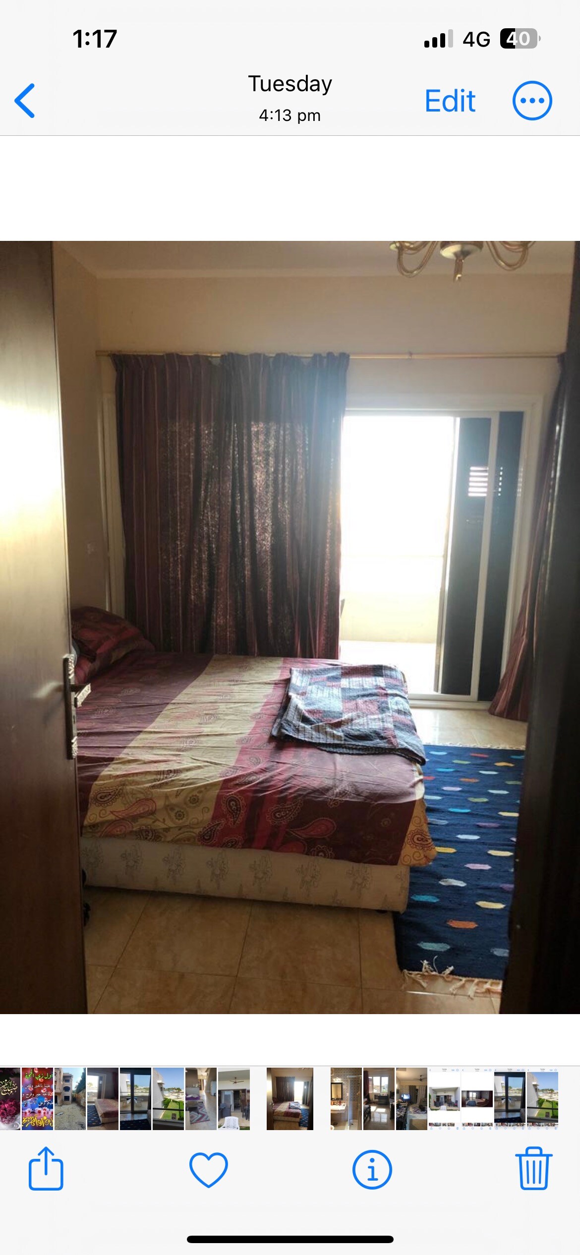 A cozy bedroom features a spacious bed with layered bedding and decorative throws. The large window is dressed with dark curtains, allowing natural light to filter in. A colorful rug adds warmth to the tiled floor, complementing the overall inviting space.