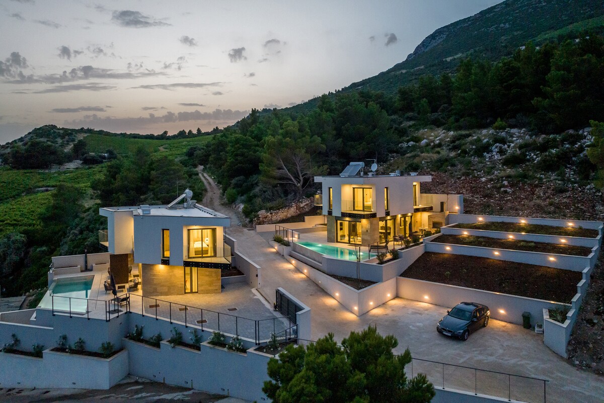 An aerial view of a modern villa complex is presented, surrounded by lush greenery and mountains. The structure features multiple levels with large windows and spacious terraces, each with pools illuminated by soft lighting. A paved pathway leads up to the villa from the driveway.