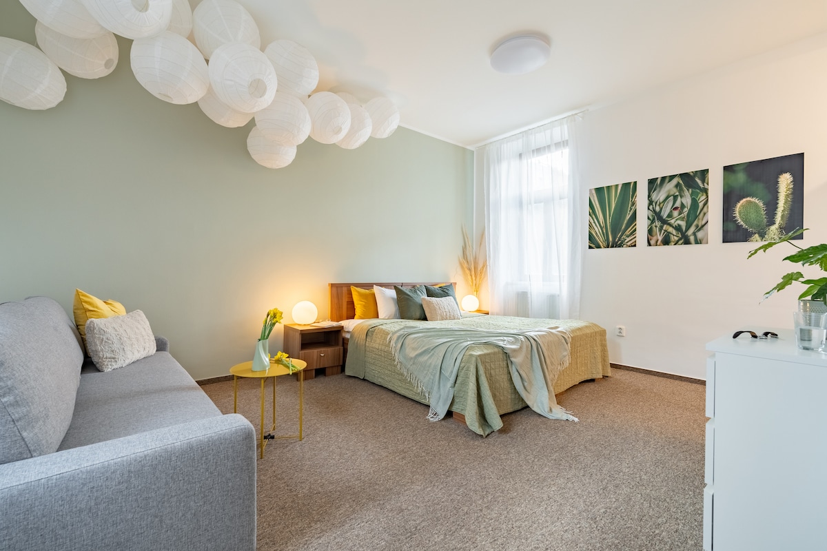 The room features a spacious bed with a light green coverlet, complemented by soft lighting from round paper lanterns. A cozy seating area is arranged with a gray sofa, and nature-themed wall art decorates one side. A plant adds a touch of greenery to the space.