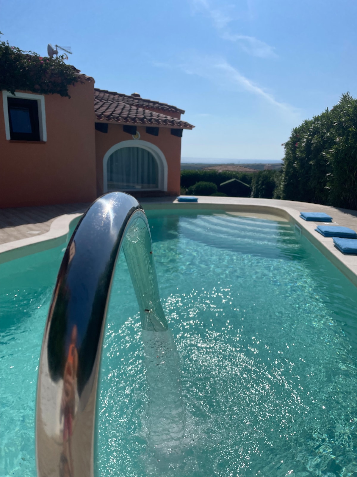 A glistening swimming pool is visible, featuring a sleek water feature that cascades gracefully. The surrounding patio is lined with sun loungers facing a scenic view of lush greenery and distant hills beneath a clear blue sky.
