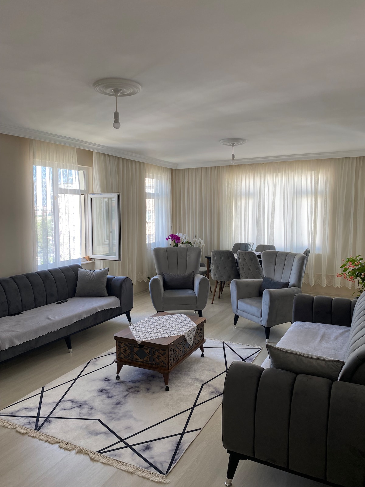A comfortable living area is displayed, featuring two gray sofas and two armchairs arranged around a low coffee table. A decorative rug lies beneath, while sunlight gently filters through sheer curtains at the windows. A dining table is positioned in the background.