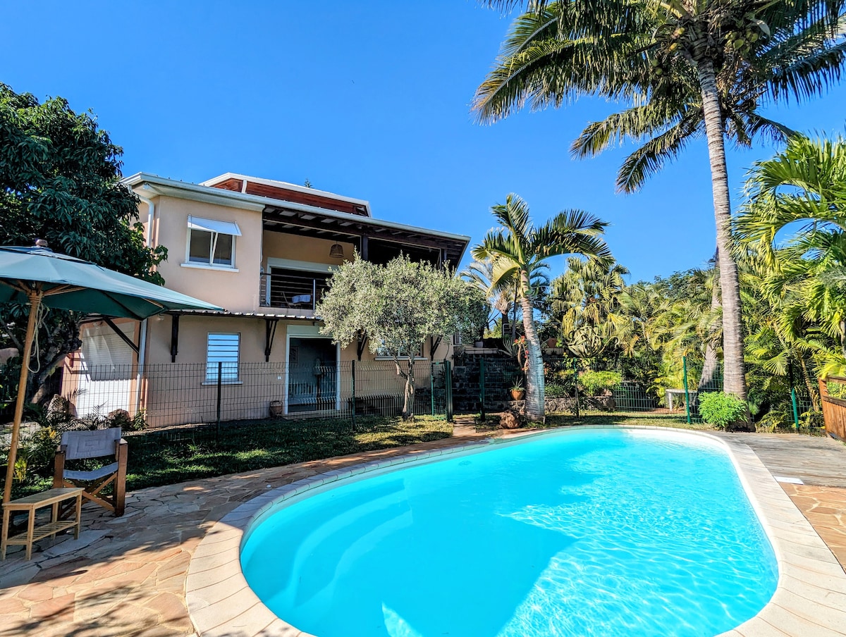 A serene outdoor space is displayed, featuring a private swimming pool surrounded by lush greenery and palm trees. The villa, with multiple levels, stands in the background, offering a sense of tropical relaxation. An umbrella provides shade beside a seating area.